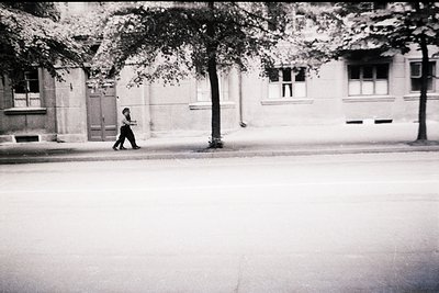 Vintage black-and-white street scene featuring a lone pedestrian crossing an empty roadway. Mid-century urban architecture wi...