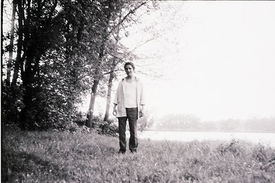 Black-and-white portrait of a lone individual standing in a misty, wooded landscape by a body of water. Mid-century style clo...