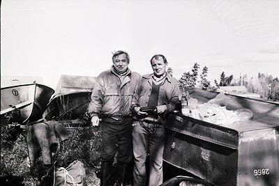 Two men pose outdoors beside a vintage truck (1960s-70s) in rural setting. One wears a quilted jacket, the other a button-dow...