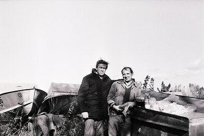 Two men pose outdoors beside a wooden boat and a trailer loaded with fish, likely in a coastal or lakeside setting. Mid-20th ...