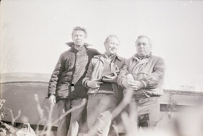Black-and-white photo of three men in outdoor industrial setting, mid-20th century. Left: man in dark jacket, hands on hips. ...