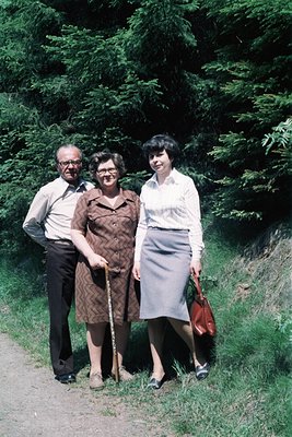 Three individuals pose outdoors in mid-20th century attire—man in suit, woman in patterned dress with cane, woman in white bl...