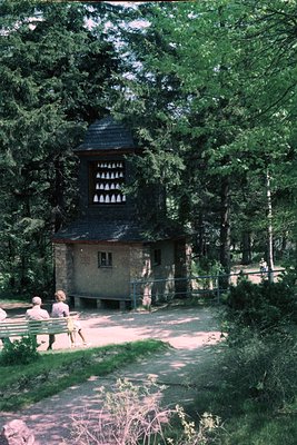 Wooden guard tower with slatted ventilation openings set in a forested area, surrounded by dense pine trees. Two individuals ...