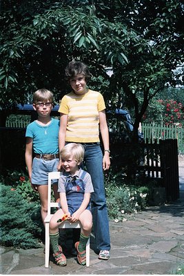 Three children pose outdoors in a lush garden setting, likely mid-1970s. The boy on the stool wears a patterned shirt, knee-h...