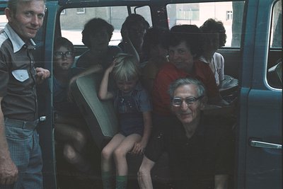 Family group shot inside a vintage van, likely 1970s–1980s. Seven individuals—adults and children—packed closely, some seated...