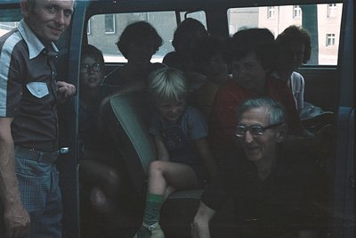 Vintage group photo inside a bus, likely 1970s Eastern Europe. A man in a striped shirt stands by the open door, while others...