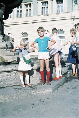 Vintage 1960s street scene with three children posing near a classical statue. Girl in white dress, knee-high socks, and red ...