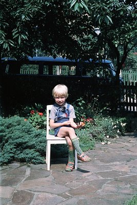 Blond-haired child in 1970s-style overalls and knee-high socks sits on a white metal chair in a lush garden. Blue double-deck...