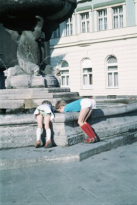 Two children play near a stone fountain in an urban plaza, 1960s-70s. Boy in red boots and striped shirt leans forward; girl ...