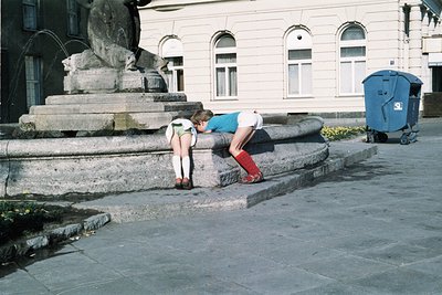 Two individuals kneeling on stone steps beside a fountain statue in an urban plaza, likely 1980s–1990s. Classic Soviet-era ar...