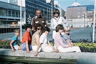 Group portrait in an urban plaza, likely 1970s. Seven individuals—two adults and five children—pose on a concrete ledge with ...
