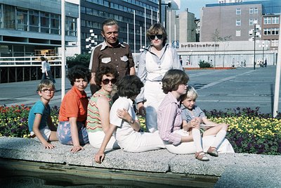 Family portrait in an urban plaza, 1970s. Seven individuals pose on concrete steps with mid-rise buildings and a fence in the...