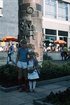Two children pose in 1970s Eastern Bloc-style clothing beside a Soviet-era monument featuring reliefs of workers and industri...