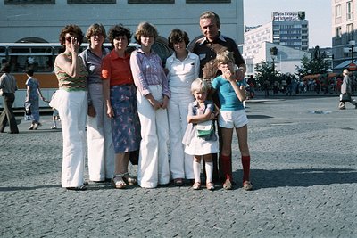 Family group shot in 1970s European city square, likely or . Adults in coordinated retro swimwear and patterned dresses; chil...