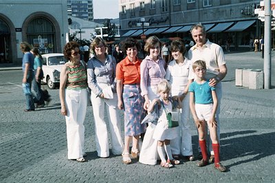 Family portrait in a European urban square, likely 1970s. Six adults and two children pose in retro summer attire—bright flor...