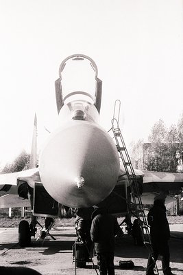 Military jet (likely **MiG-29**) with open canopy, showcasing twin-engine, afterburner-equipped design. Ground crew in dark u...