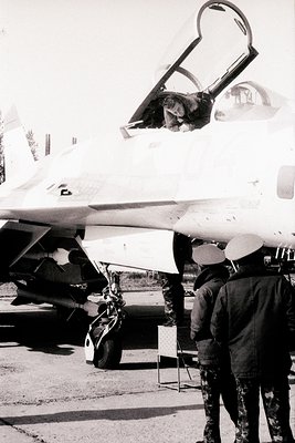 Military jet cockpit open, revealing pilot seated inside. Two uniformed personnel in camouflage inspect or prepare aircraft. ...
