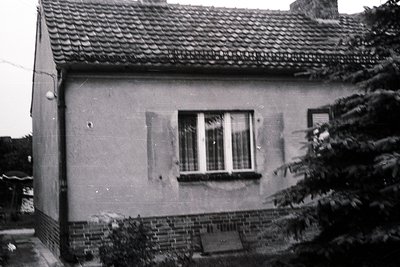 Single-story brick-and-plaster house with tiled roof, showing wear. Symmetrical window with divided panes and closed blinds. ...