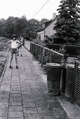 1970s-era concrete pathway with metal fencing and large trash bins, likely Eastern European residential area. A shirtless ind...