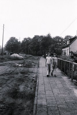 Two men in 1970s-style clothing walk along a paved riverside path flanked by a metal railing. Overgrown vegetation and a rock...