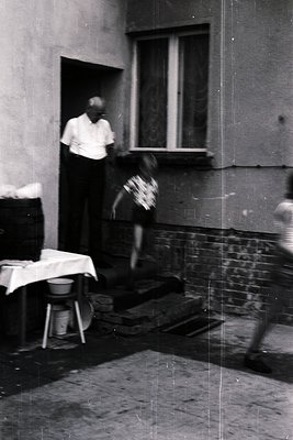 Vintage black-and-white photo of an elderly man in a doorway, holding a child mid-jump in a dimly lit courtyard. Brick wall, ...