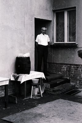 Mid-20th century man in light button-down shirt leans against a weathered brick building’s doorway. A large woven basket and ...