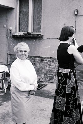 Vintage black-and-white photo of an elderly woman in a white apron standing beside a younger woman in a patterned dress, like...