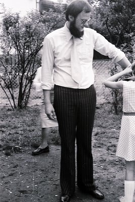 Portrait of a man in a loose-collared button-up shirt and pinstripe trousers, standing outdoors beside a white mesh fence. Hi...