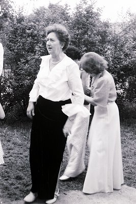 Mid-20th century outdoor portrait: two women in formal attire—one in a fitted blouse and black trousers, the other in a white...