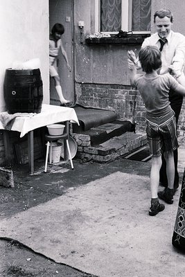 Mid-20th century courtyard scene: two boys play near a barrel and stools, one in plaid shorts, the other in briefs. Adult in ...
