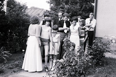 Group portrait in a residential garden, 1970s. Nine individuals pose outdoors near a brick house and overgrown shrubs, reflec...