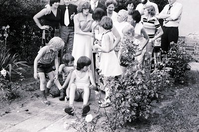 A lively 1970s outdoor gathering in a lush garden. Adults in casual summer attire (dresses, shorts, striped shirts) and child...