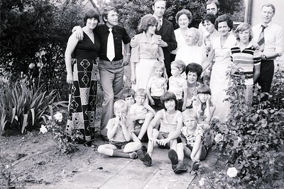 Family gathering in a lush garden, 1970s. Adults in formal attire (ties, dresses) stand behind seated children in casual summ...