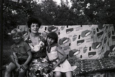 A black-and-white family portrait from the 1960s–1970s, featuring a woman and two children seated on patterned fabric. The wo...