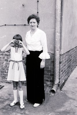 A woman in a 1960s-style blouse and pencil skirt teaches a young girl to use a vintage camera in an urban courtyard. The bric...