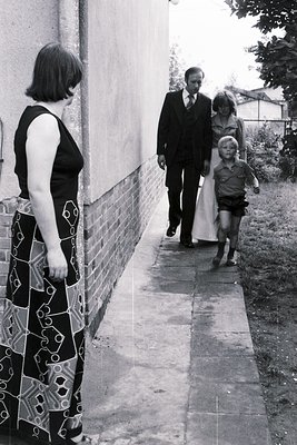 A black-and-white snapshot of a mid-20th-century family moment: a woman in a geometric-patterned skirt and sleeveless top sta...
