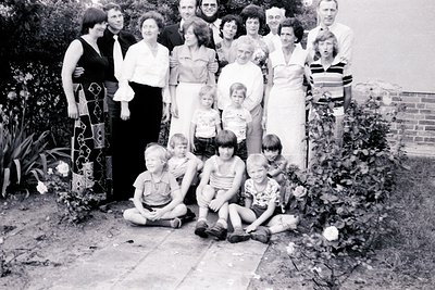 Family portrait in a garden setting, likely mid-20th century (1950s–1970s). Nine adults and six children pose outdoors on pav...