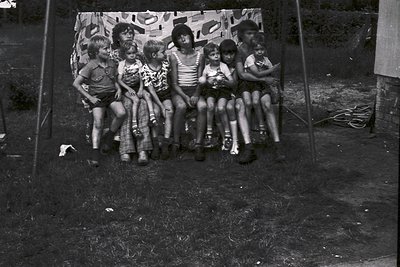 Group of children posing on a makeshift wooden bench in an outdoor setting, likely 1960s–1970s. Casual summer attire includes...
