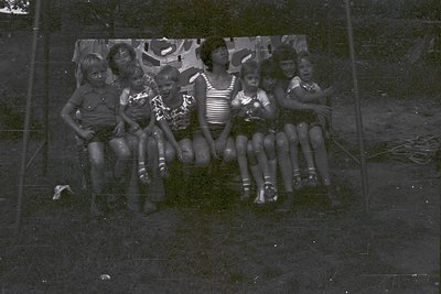 Vintage black-and-white group portrait of seven children seated on a chain-link fence, mid-20th century. Boys wear shorts and...
