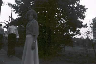 Vintage black-and-white photo of a woman in a knee-length dress and blouse, standing outdoors under leafy trees. Her hair is ...