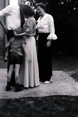 Vintage black-and-white photo of a family moment: a woman in a long, flowing dress lifts a young boy in a plaid shirt, while ...