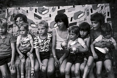 Black-and-white group portrait of seven children and two adults seated on a camouflage-patterned wall. Mid-20th century cloth...