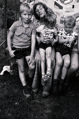 Three children in mid-20th-century clothing—plaid shorts, knee-high socks, and short-sleeve shirts—sit closely on grassy grou...
