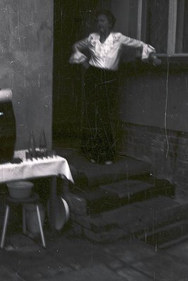 Blurred black-and-white interior shot of a man in mid-motion, likely playing a piano, reflected in a foggy window. Visible th...