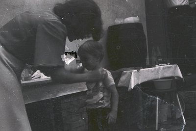 A mid-20th-century kitchen scene: an adult and child at a metal countertop, blurred vintage filter. The adult leans over a ch...