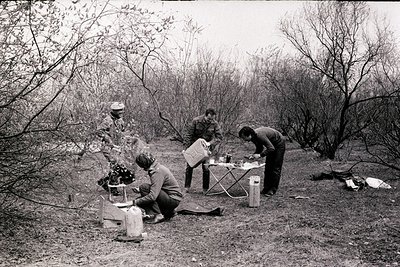 Three individuals in 1960s-70s outdoor camping gear prepare a meal by a portable stove and folding table in a wooded area. On...