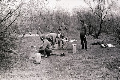 Three men in 1970s outdoor camping attire—folding chairs, camping stove, and backpacks—set up near a wooded area. Mid-century...