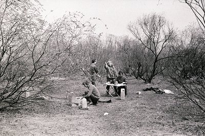 Three individuals gather around a makeshift campsite in a wooded area, likely mid-20th century. A man kneels by a small fire ...