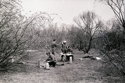 Three individuals in military uniforms sit on a makeshift wooden bench in a wooded area, likely mid-20th century. One stands,...