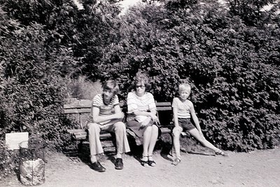 Three individuals pose on a wooden bench in a lush, green park setting, mid-20th century. Adult wears a short-sleeve blouse a...